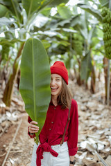 Wall Mural - Portrait of a cute woman dressed in red hat and shirt standing with a green banana leaf on the plantation. Vegetation for wellness concept