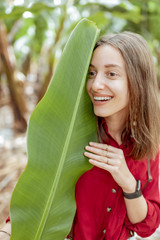 Wall Mural - Facial portrait of a cute smiling woman hiding behind a banana leaf on the plantation. Vegetation for wellness and cosmetics concept