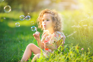 Cute girl blows soap bubbles while sitting on the grass in the park at sunset. Children's fun..