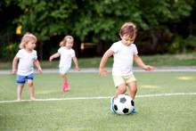 Football Game: Team Of Toddlers Playing Soccer On Green Field: Three Children, Two Boys (one Is Barefoot) And Girl Playing At Stadium, Smiling Little Boy Dribbling Ball Is Running In Front Place