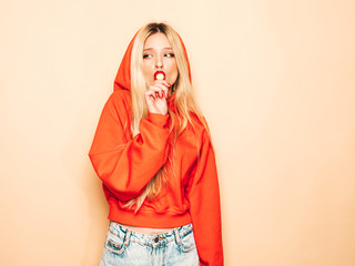 Portrait of young beautiful hipster bad girl in trendy red summer hoodie and earring in her nose.Sexy carefree smiling blond woman posing in studio.Positive model licking round sugar candy