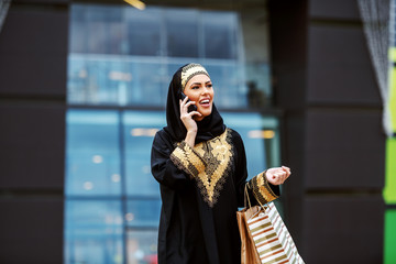 Stunning attractive positive smiling muslim woman in traditional wear standing in front of shopping mall with shopping bags in hands and calling taxi.