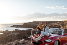 Young Lovely Couple Enjoying Landscapes, Sitting Together On A Car Hood, Traveling By Car On The Rocky Ocean Coast. Carefree Lifestyle, Love And Travel Concept