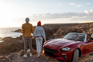 Couple enjoying beautiful views on the ocean, standing together near the car on the rocky coast. Carefree lifestyle, love and travel concept