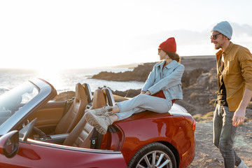 Young joyful couple having fun on the rocky coast while traveling by car on a sunset. Carefree lifestyle, love and travel concept