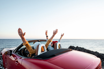 Couple driving convertible car, traveling near the ocean on a sunset, view from the backside. Happy vacations and traveling by car concept