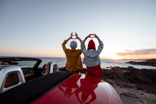 Couple Enjoying Beautiful Views On The Ocean, Standing Together Near The Car On The Rocky Coast, Showing With Hands Heart Shape. Carefree Lifestyle, Love And Travel Concept