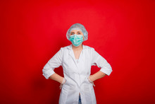 A Woman Doctor In A White Coat, Mask, Rubber Gloves And A Medical Cap Is Standing On A Red Background Holding His Hands On His Belt.