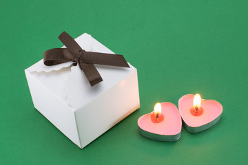 Two burning pink candles in the form of a heart and white, paper, square, gift box with a brown ribbon on a green background. Little box for the holiday.