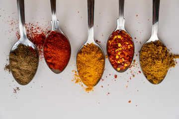 Top view of spoons filled with different spices on a white background. Ground pepper, smoked paprika, turmeric, chilly flakes and ground cumin.