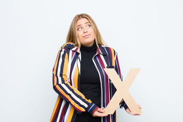 young pretty blonde woman confused, doubtful, thinking, holding the letter X of the alphabet to form a word or a sentence.