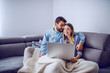 © Dusan Petkovic - Cheerful cute caucasian couple sitting on sofa covered with blanket, snuggling and using laptop for internet surf. Living room interior.