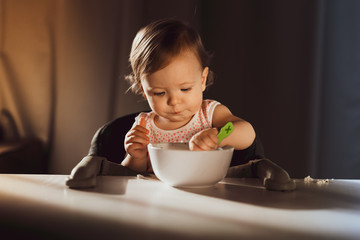 Wall Mural - Nine-month-old smiling baby girl in pink bandage on her head sits at white table in highchair and eats herself with spoon from bowl. Blurred background. Healthy eating for kids. Child's nutrition.