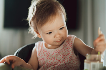 Wall Mural - A nine-month-old smiling baby girl sits at a white table in a highchair and eats herself with a spoon from a bowl. Dark background. Healthy eating for kids. Child's nutrition.