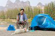 © chayathon2000 - Asian young man in blue shirt with cute puppy dog camping on the lake hill mountain view happy and enjoy life