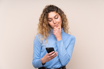 Wall Mural - Young blonde woman with curly hair wearing a turtleneck sweater isolated on beige background thinking and sending a message