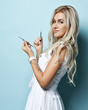 Smiling sexy blond cosmetologist in medical gown is posing sideways to us holding cosmetology tools over blue background