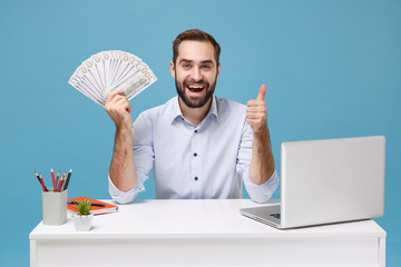 Joyful young man in shirt work at desk with pc laptop isolated on blue background. Achievement business career concept. Mock up copy space. Hold fan of cash money in dollar banknotes showing thumb up.