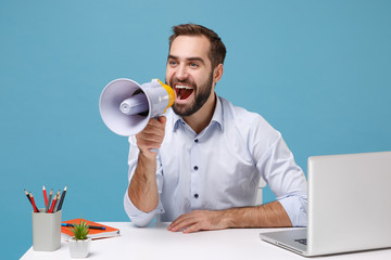 Funny young bearded man in light shirt sit work at desk with pc laptop isolated on pastel blue background. Achievement business career concept. Mock up copy space. Scream in megaphone, looking aside.