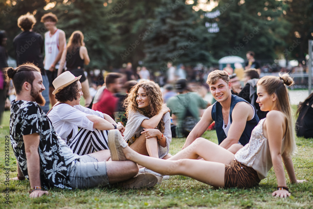 Group of young friends sitting on ground at summer festival. - obrazy, fototapety, plakaty 