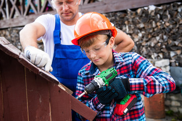 Cute boy in a construction helmet and glasses with a screwdriver in his hands building wooden dog house with his father. Construction and decoration of wooden houses