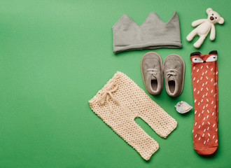 Wall Mural - Eco baby clothes and accessories concept. Knitted crown, pants, socks, bear toy and shoes on green background with blank space for text. Top view, flat lay.