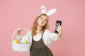 Little blonde kid girl 11-12 years old in spring dress bunny rabbit ears hold in hand cell phone wicker basket colorful eggs isolated on pastel pink background Childhood lifestyle Happy Easter concept