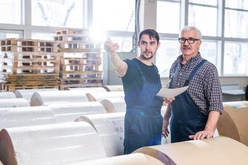 Wall Mural - Senior and young warehouse workers of printing plant standing among paper rolls and discussing delivery