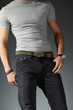 Cropped shot of a man in black jeans and a gray t-shirt on the gray background. A khaki woven belt with brown leather fixings is fitted with a steel buckle and a leather loop.