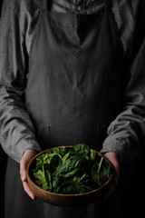 Wall Mural - wooden plate with fresh spinach leaves in the hands of a woman dressed in gray linen clothes. closeup