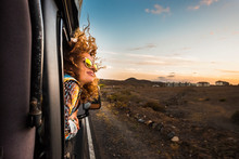 Beautiful Caucasian Young Woman Travel Outside The Car With Wind In The Curly Hair, Motion And Movement On The Road Discovering New Places During A Nice Sunset, Enjoy And Joyful Freedom Concept