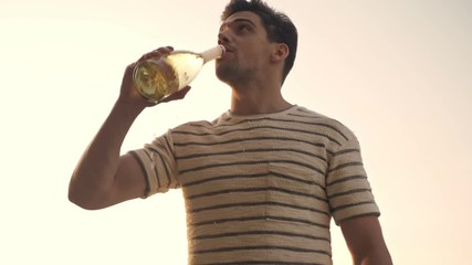 Canvas Print - A bottom view of a handsome man is opening the bottle of champagne and drinking it outdoors in summer