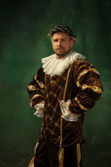 Posing thoughtful. Portrait of medieval young man in vintage clothing standing on dark background. Male model as a duke, prince, royal person. Concept of comparison of eras, modern, fashion.