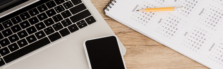 Panoramic shot of smartphone on laptop with calendar and pencil on wooden background