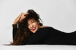 Happy laughing young woman with long curly brunette hair in black tight dress is lying on the floor