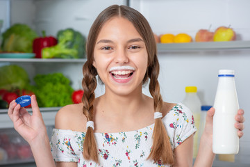 Beautiful young teen girl holding bottle of milk and drinks while standing near open fridge in kitchen at home. Portrait of pretty child choosing food in refrigerator full of healthy products.