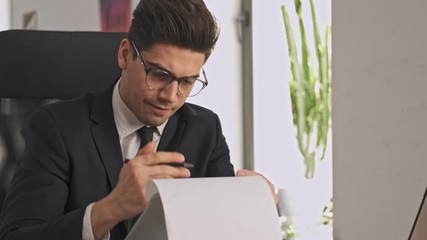 Poster - Concentrated businessman in formal suit and eyeglasses studying a documents with clipboard while sitting by the table in office