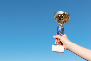 Children's hand holds a trophy cup. Championship cup and blue sky in the background. The joy of the winner. Award and victory concept. Background. Space for text.