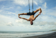 Outdoors Portrait Of Young Attractive And Athletic Man Practicing Aero Yoga Workoutat The Beach Hanging From Rope Swiing Above The Sea Training Body Balance And Flexibility
