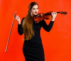 Portrait of a pretty brunette musician girl with a smile in a black dress on a red background holds a violin in her hands