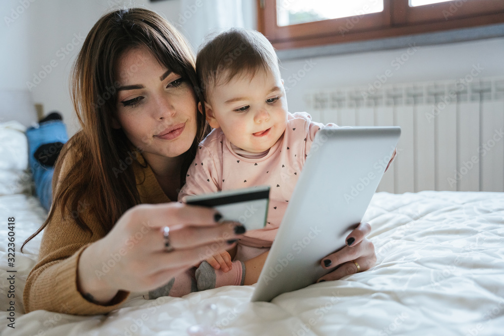 Portrait of a beautiful young mom with a baby girl lying on the bed in a moment of intimacy while buying products online with the tablet and holding her credit card in hand - Millennial with daughter - obrazy, fototapety, plakaty 
