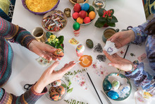 White Table With Colored Easter Eggs In Baskets And Bowls On It, Along With A Lot Of Arts And Crafts Tools. Two Women Are Painting Some Of The Eggs By Hand Using Decoupage Technique.   