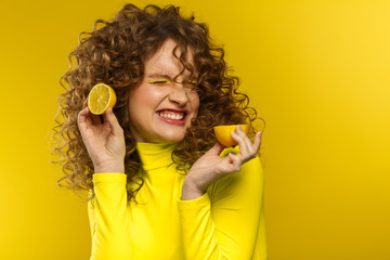 Young beautiful smiling women with curly hair tasting sour lemon halves emotionally. Funny girl with wavy hairstyle and bright make up isolated over yellow background. Copy space. Yellow lifestyle.