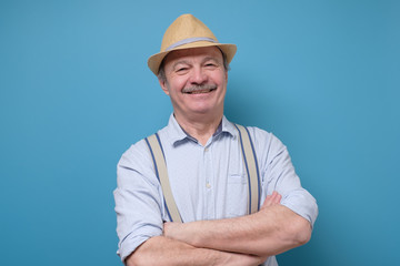 Portrait of friendly confident senior man in summer hat standing isolated over blue background. Positivre facial human emotion.