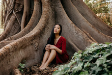 Beautiful Young Woman Wearing A Red Dress Sitting At A Tree With Large Roots