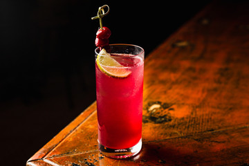 A red cocktail in a highball glass garnished with lemon and cherries