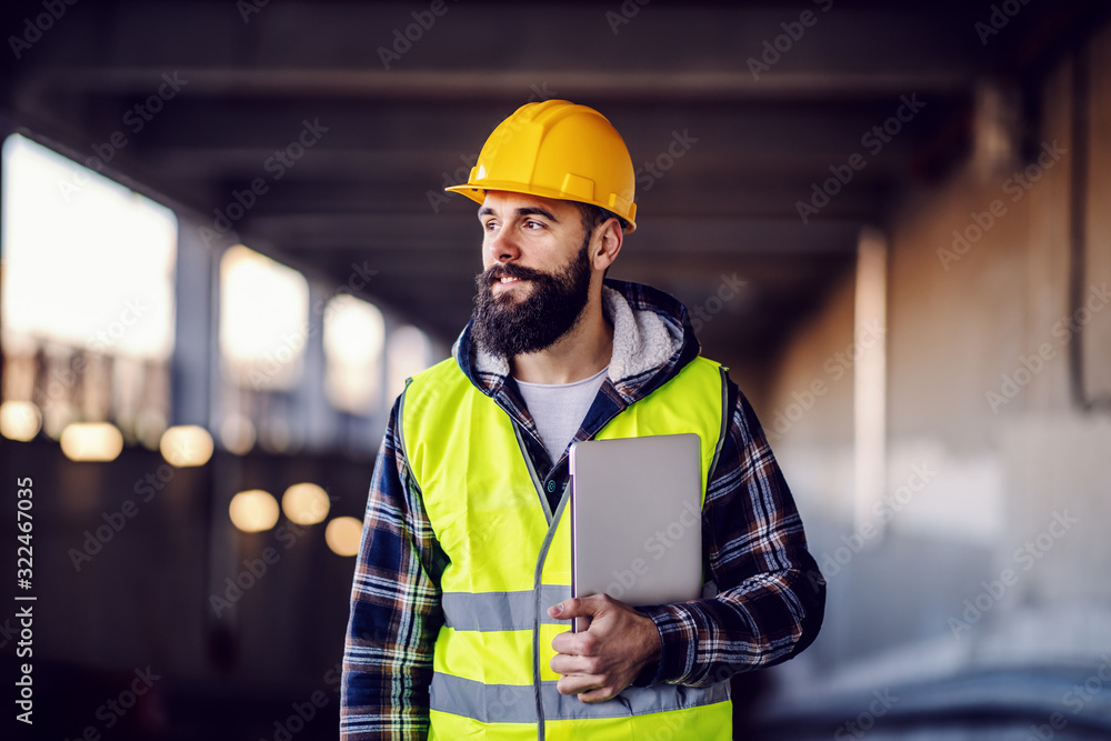Portrait of highly motivated caucasian hardworking smiling bearded supervisor with helmet on head in vest and with laptop in hands posing on construction site. - obrazy, fototapety, plakaty 