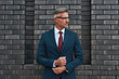 © Svitlana - Confident and stylish. Handsome mature businessman in full suit adjusting his sleeve while standing outdoors against the brick wall