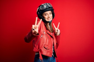 Wall Mural - Young beautiful brunette motocyclist woman wearing motorcycle helmet and red jacket smiling looking to the camera showing fingers doing victory sign. Number two.