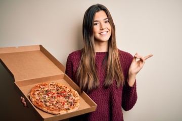 Poster - Young beautiful girl holding delivery box with Italian pizza standing over white background very happy pointing with hand and finger to the side